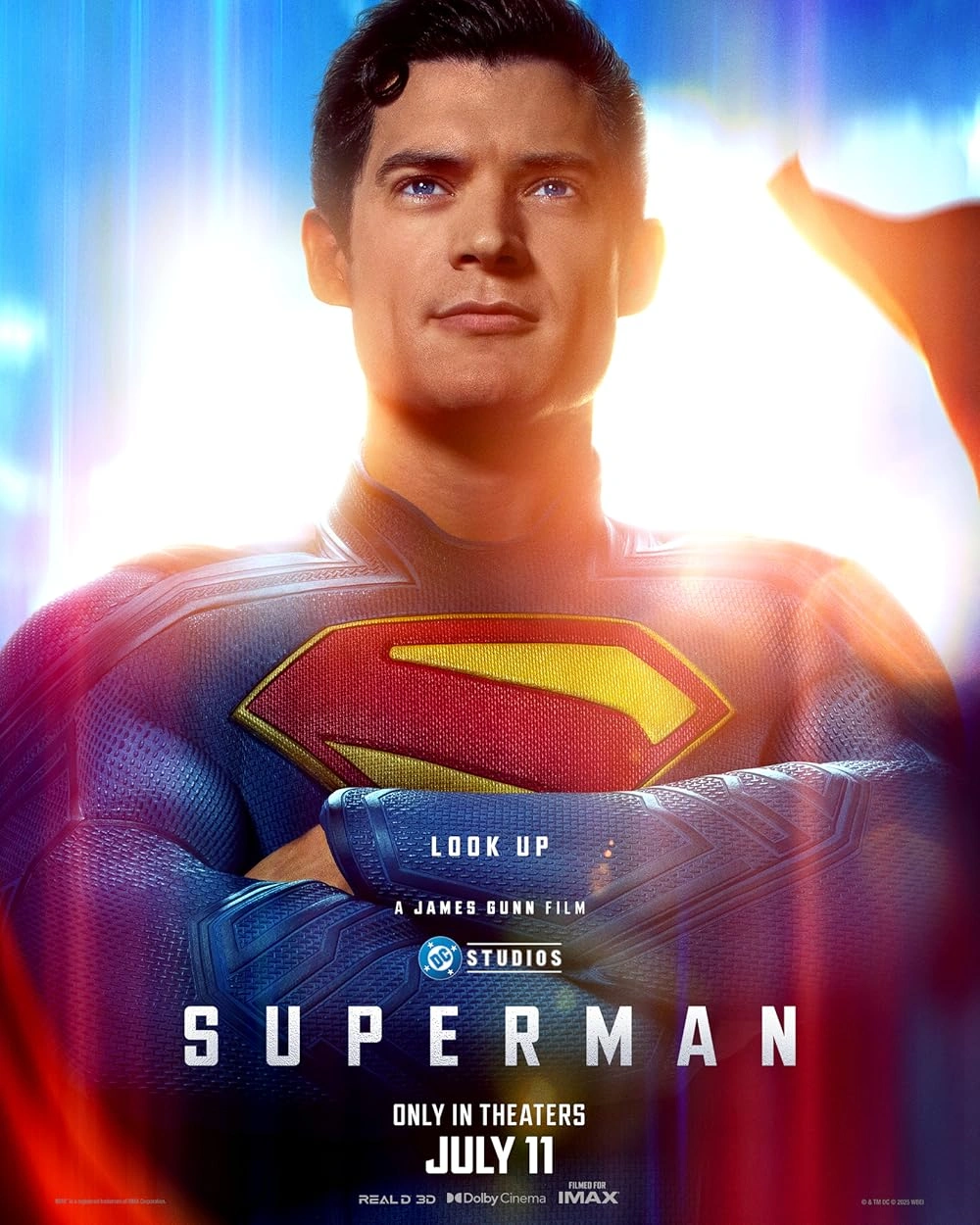 A superhero in a blue suit with a red and yellow S emblem on his chest stands with his arms crossed, looking upward. Text below reads Superman—Only in Theaters July 11. Light beams surround him.